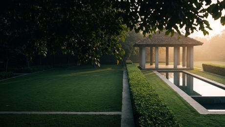 One Plot, Three Design Languages — villa estate garden at sunrise A large villa estate garden at sunrise in the dry interior of South India, with a clipped hedge and a still reflecting tank of grey limestone leading to an open pavilion, seen from beneath a shaded tree canopy
