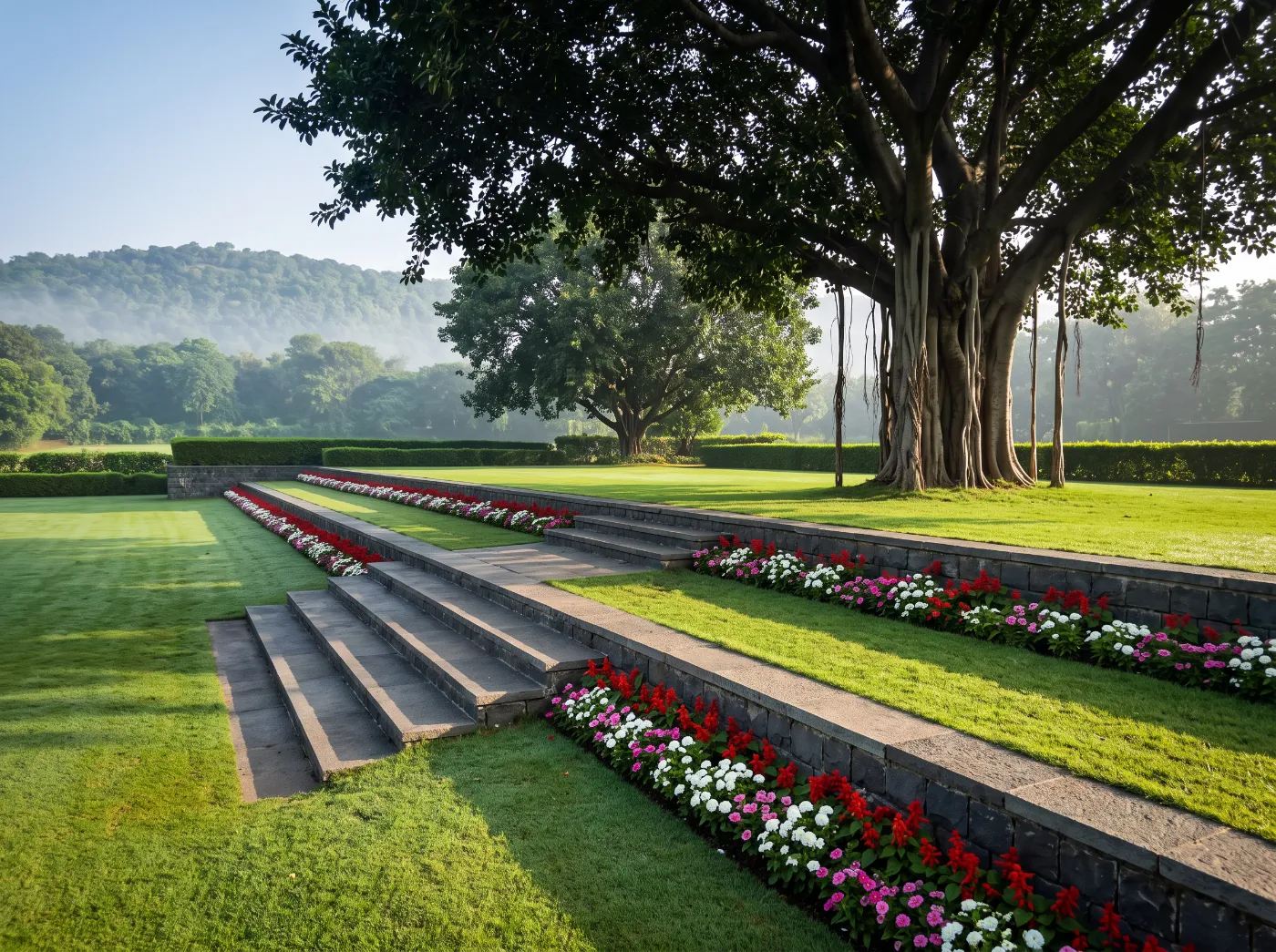 Basalt steps descending between formal lawn terraces, continuous bands of scarlet and white flowers, beneath a mature banyan with aerial roots